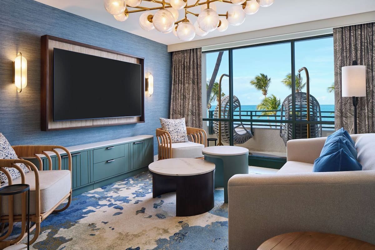A modern living room with a large flat-screen TV, comfortable seating, a round coffee table, and a view of the ocean.