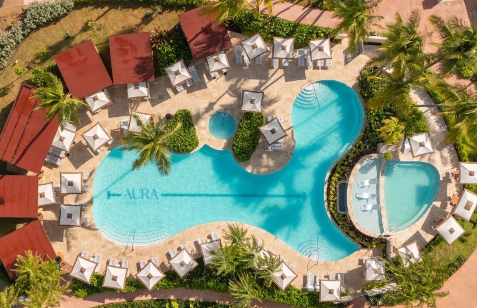 An aerial view of a resort with a large central pool, multiple sun loungers, and palm trees.