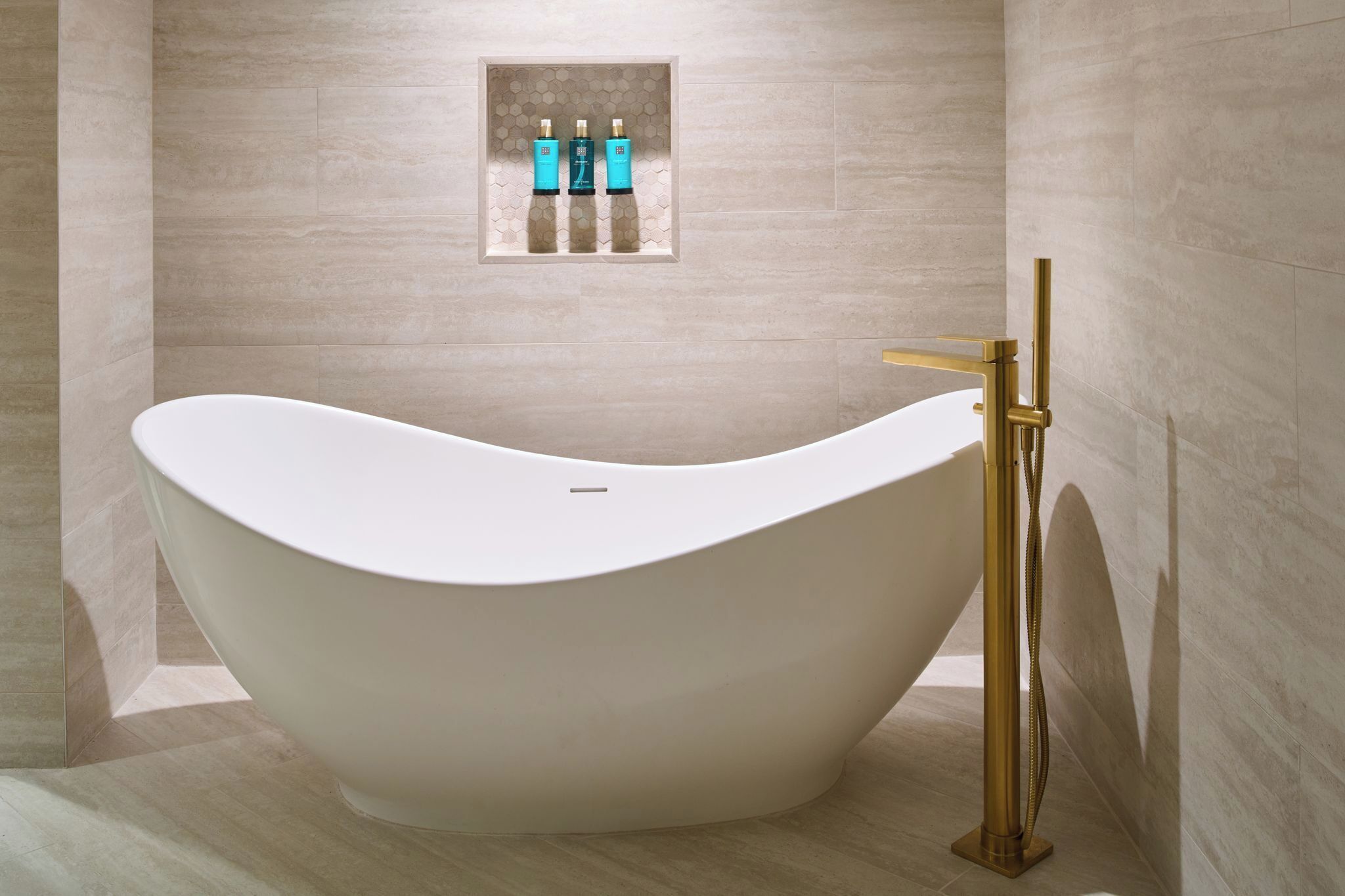 A freestanding bathtub in a modern bathroom with a hexagonal tile wall and a gold faucet.
