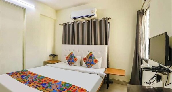 A hotel room with a bed, a bedside table, a television, and an air conditioning unit.