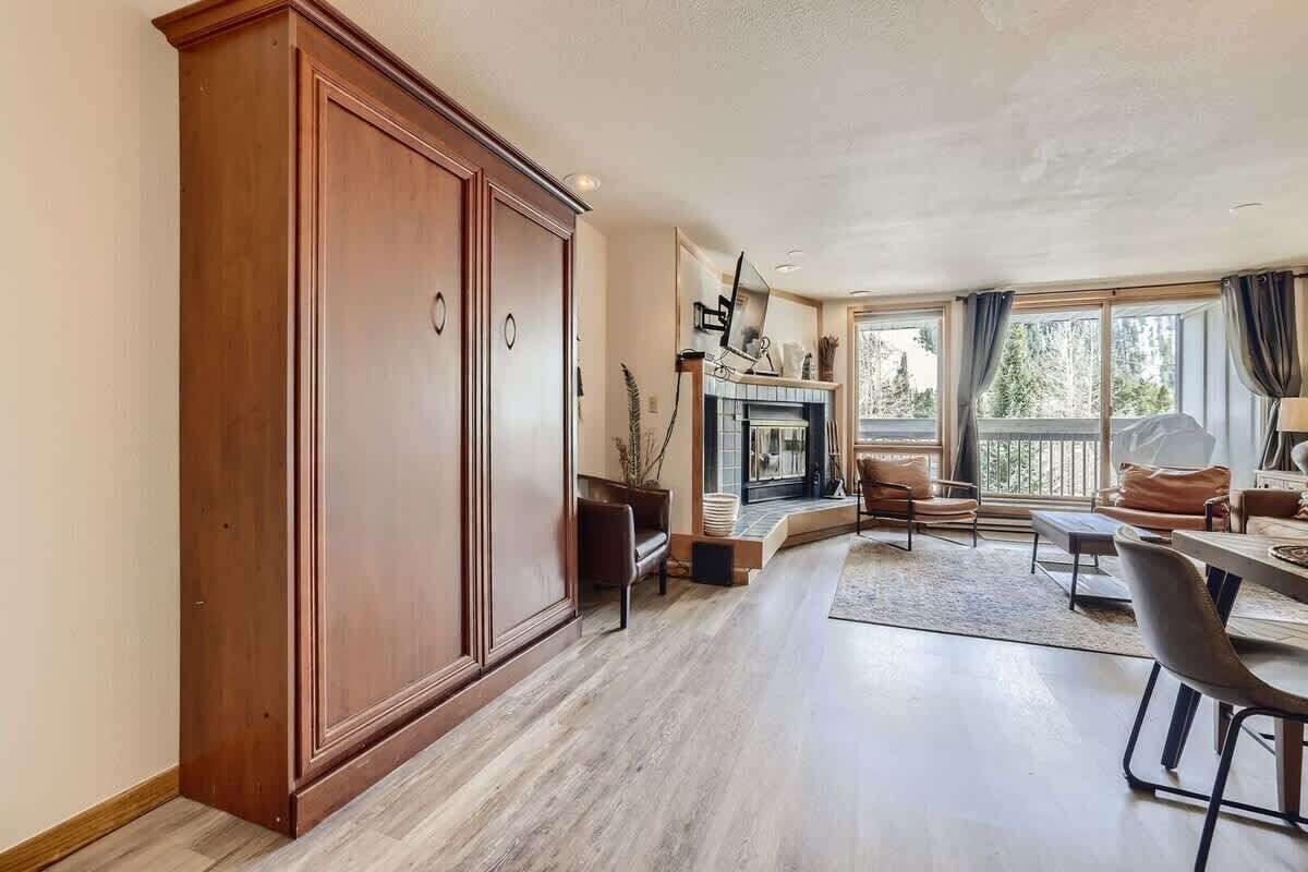 A spacious living room with a large wooden cabinet, a fireplace, and a view of the outdoors through large windows.