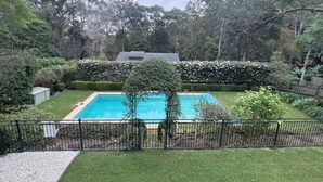 A rectangular swimming pool surrounded by a black metal fence, with a hedge in the background and trees around.