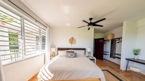 Ein Schlafzimmer mit einem Bett, Deckenventilator, Nachttisch, Ablagefläche an der Wand und einem Fenster mit Jalousien.