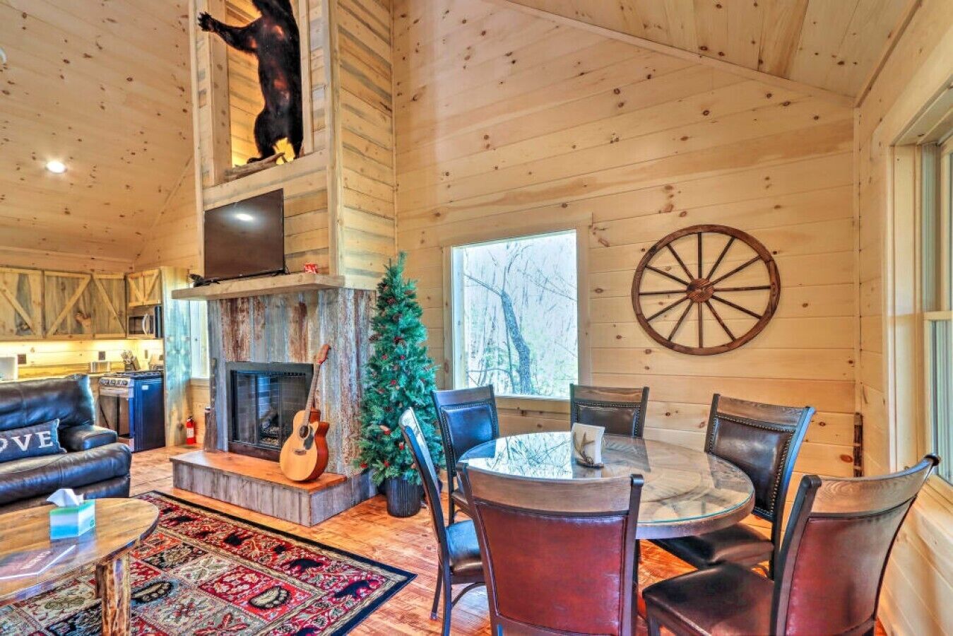 Een houten cabin interieur met een open haard, een kerstboom, een eettafel met stoelen en een groot houten wiel aan de muur.