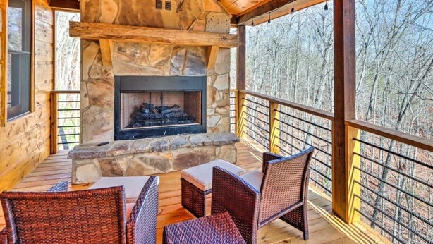 A stone fireplace with a fire, wicker chairs, and a wooden deck with a railing. - Luxury Mineral Bluff Cabin with Hot Tub and Decks (Mineral Bluff)