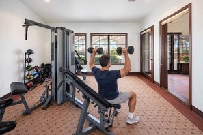 Un homme effectue un exercice de développé couché avec des haltères, assis sur un banc, dans une salle de sport équipée de divers appareils et dotée d’un tapis à motifs.