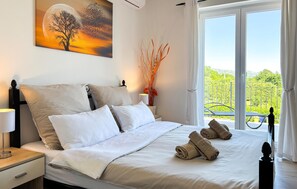 A hotel room with a bed, pillows, a nightstand with a lamp, a painting on the wall, a vase with branches, and a balcony with a view. - Stunning home in Labin (Labin)