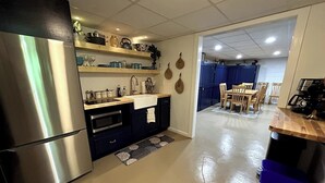 A kitchen with a stainless steel refrigerator, wooden shelves, a sink, and a dining area with chairs and a table.