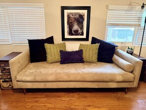 A beige sofa with three pillows, a framed picture above it, and a wooden floor.