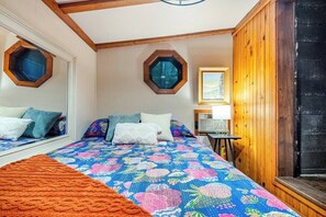 A bedroom with a large bed, two hexagonal windows, a wooden wall, and a framed picture on the wall. - New! Rustic Creekside Cabin-Hot Tub, Cozy, Private (Ansted)