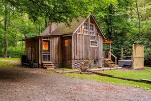 A rustic wooden cabin with a porch, a hot tub, and a fenced area. - New! Rustic Creekside Cabin-Hot Tub, Cozy, Private (Ansted)