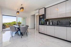 A modern kitchen with a large island, a dining area with a glass table, and a view of greenery outside. - Holiday house VILLA PATRICK with pool, jacuzzi and sauna (Kornić)