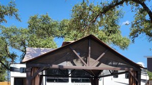Un espacio al aire libre cubierto con zona de barbacoa, mobiliario de mimbre y vista a una autocaravana blanca al fondo.