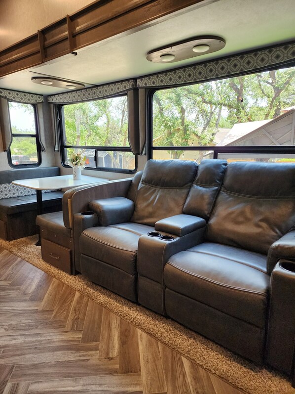 A cozy RV interior with a leather sofa, a small table with a vase, and large windows offering a view of trees. - Canyon Lake Astoria RV with 2 bedrooms and 2 bathrooms and AC (Canyon Lake)
