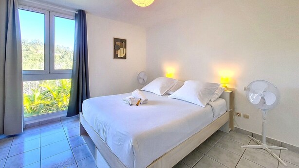 Una habitación de hotel con cama, lámparas de noche, ventilador de pared y ventana con cortinas.