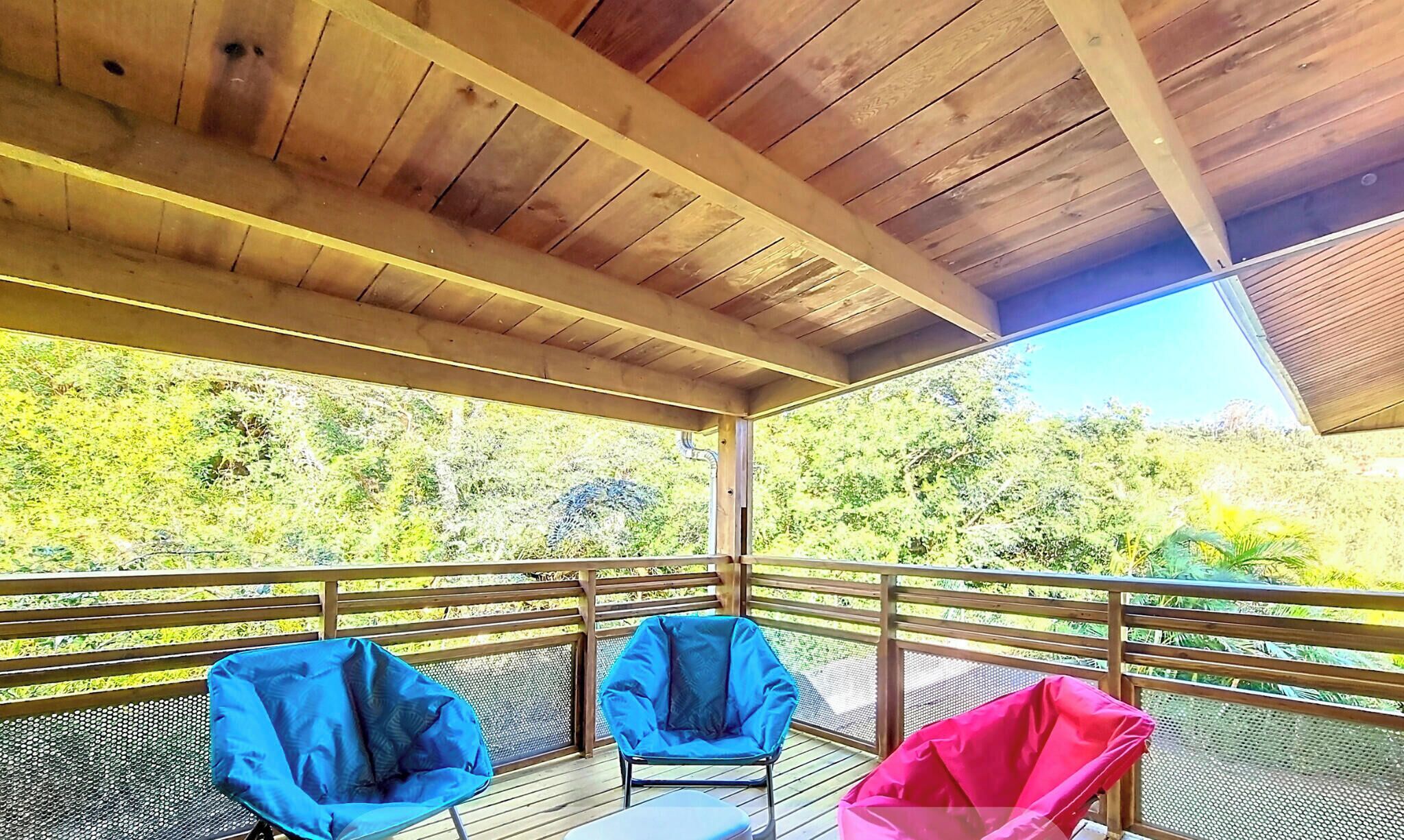 A covered outdoor deck with a wooden ceiling and metal railing. There are three chairs, two blue and one pink, on the deck.