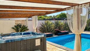 Uma banheira de hidromassagem e uma piscina sob uma pérgola de madeira, com vista para uma parede de pedra e vegetação.