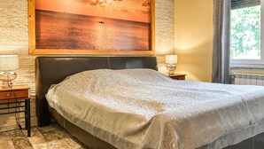 Quarto com cama, mesa de cabeceira, luminária, pintura de um pôr do sol sobre a água e janela com cortinas.