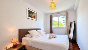 Una cama bien hecha con sábanas blancas, una mesita de noche con lámpara, un cuadro en la pared y una ventana con cortinas.
