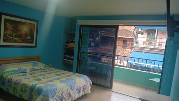 A bedroom with a bed, a desk, a chair, a window with a view of buildings, and a painting on the wall.