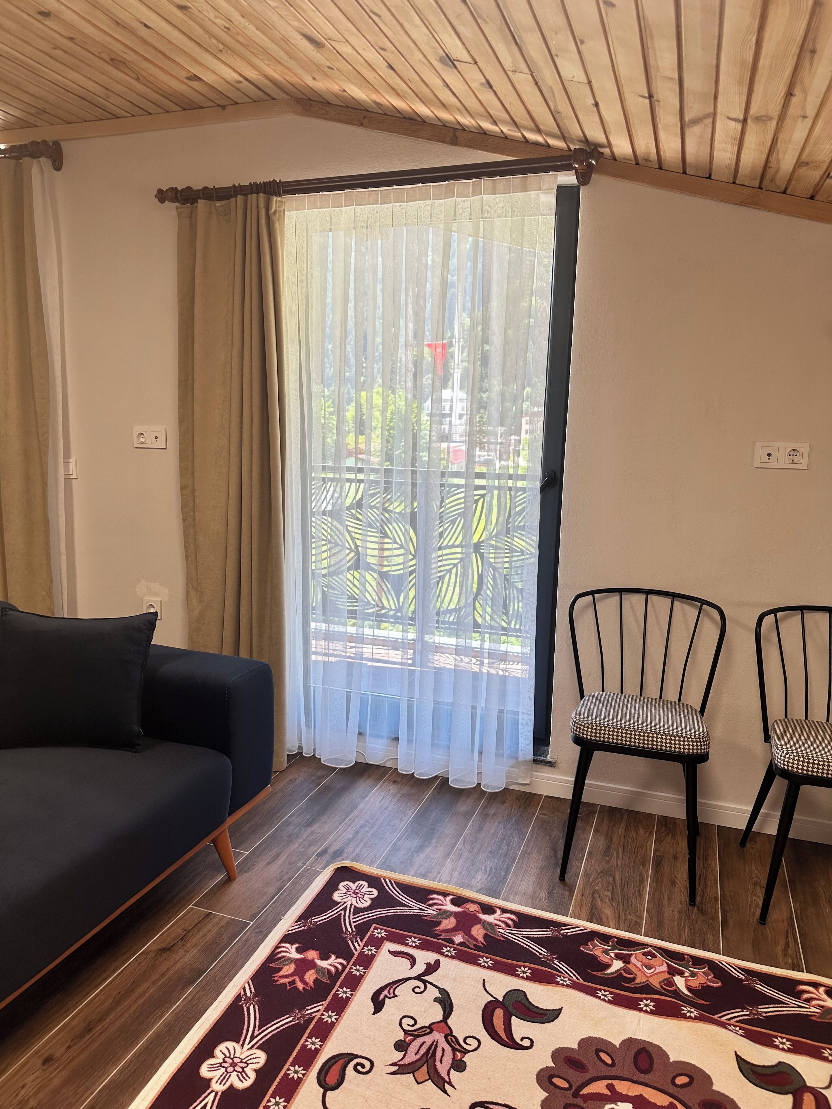 Une pièce avec un canapé foncé, deux fauteuils, un tapis à motifs, un plafond en bois et une fenêtre ornée de rideaux légers.