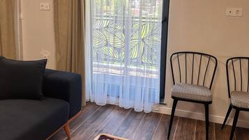 Une pièce avec un canapé foncé, deux fauteuils, un tapis à motifs, un plafond en bois et une fenêtre ornée de rideaux légers.