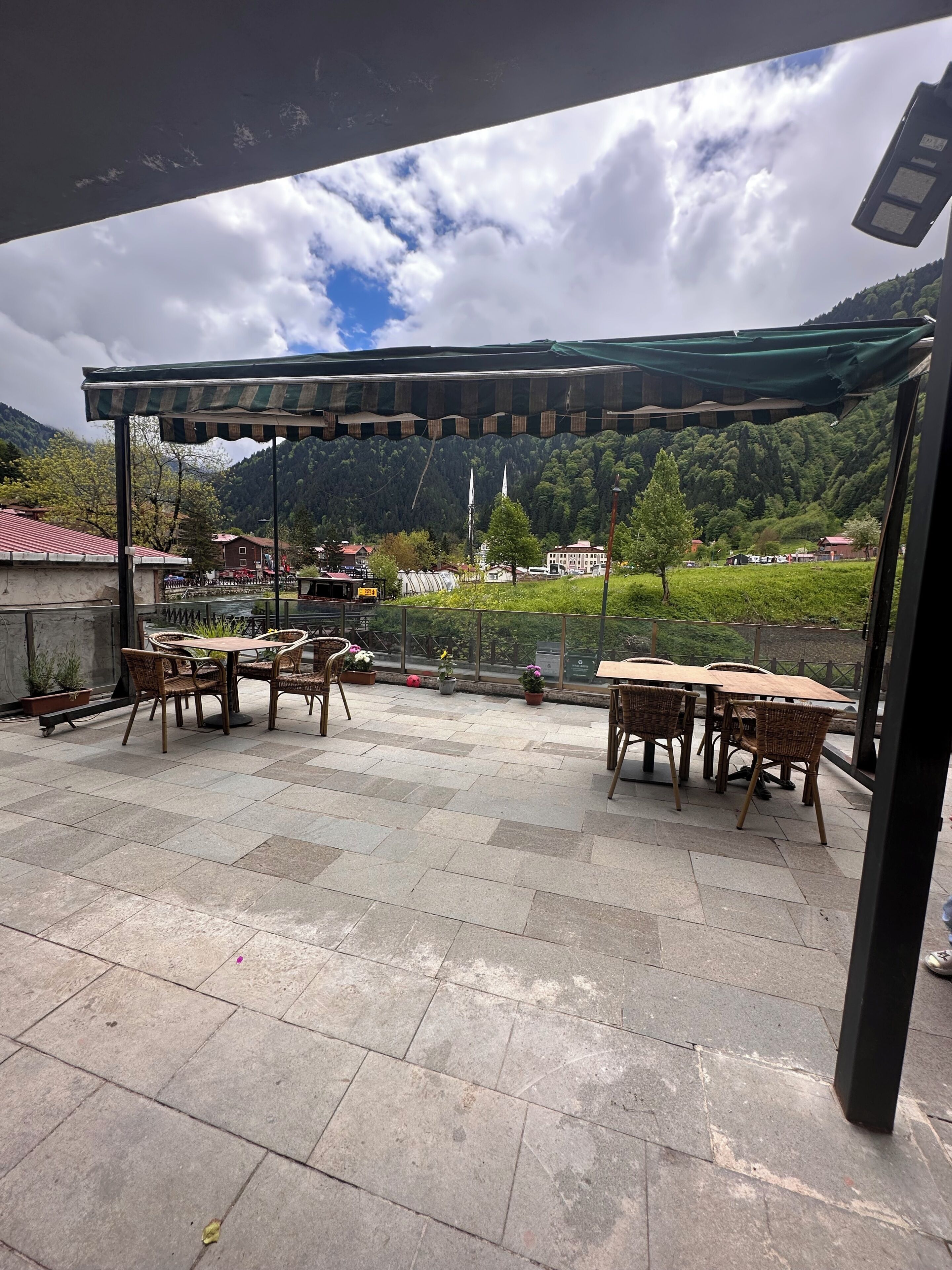 Een terras buiten met tafels en stoelen, een pergola en uitzicht op een bergachtig landschap.