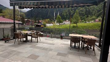Een terras buiten met tafels en stoelen, een pergola en uitzicht op een bergachtig landschap.