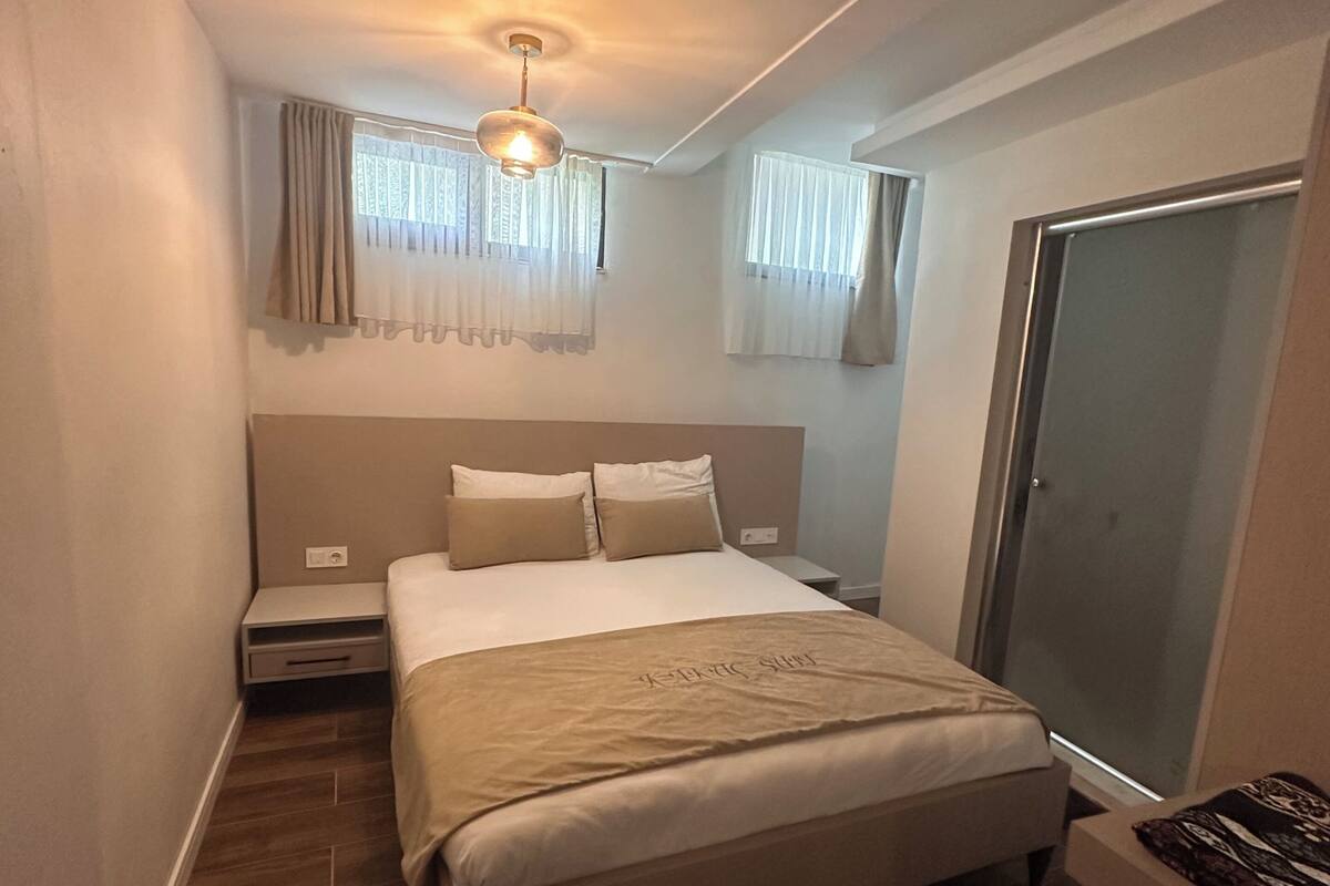 A bedroom with a bed, bedside tables, a ceiling light, a window with curtains, and a door.