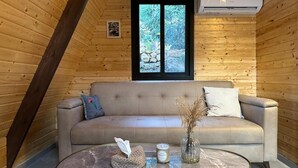 A wooden cabin interior with a sofa, a round table, and a window with a view of greenery.
