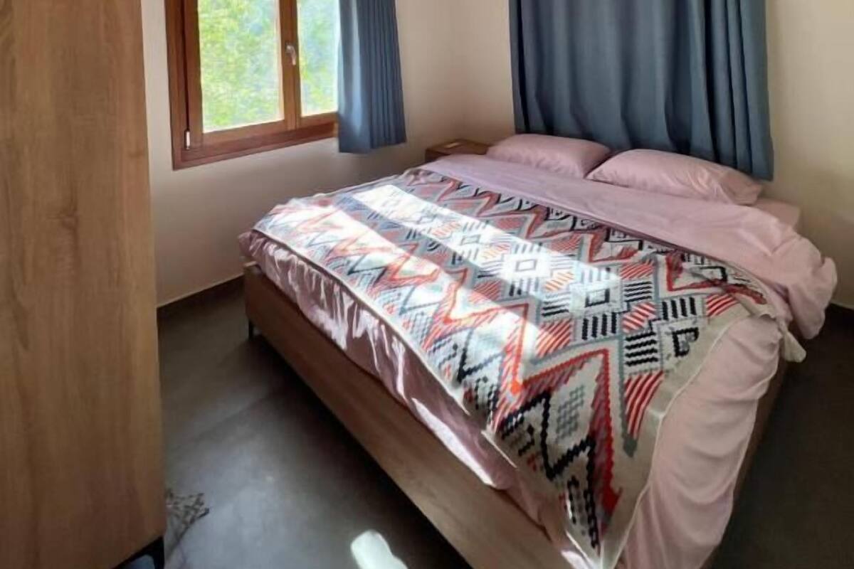 A bedroom with a sloped ceiling, a bed with patterned bedding, a window with curtains, and a wooden wardrobe.