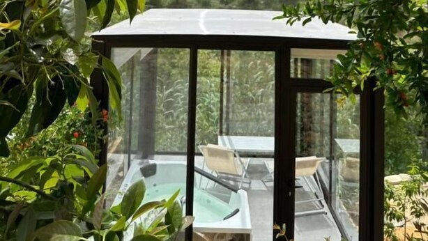 A glass-enclosed outdoor seating area with a pool, surrounded by lush greenery and a mountainous backdrop.