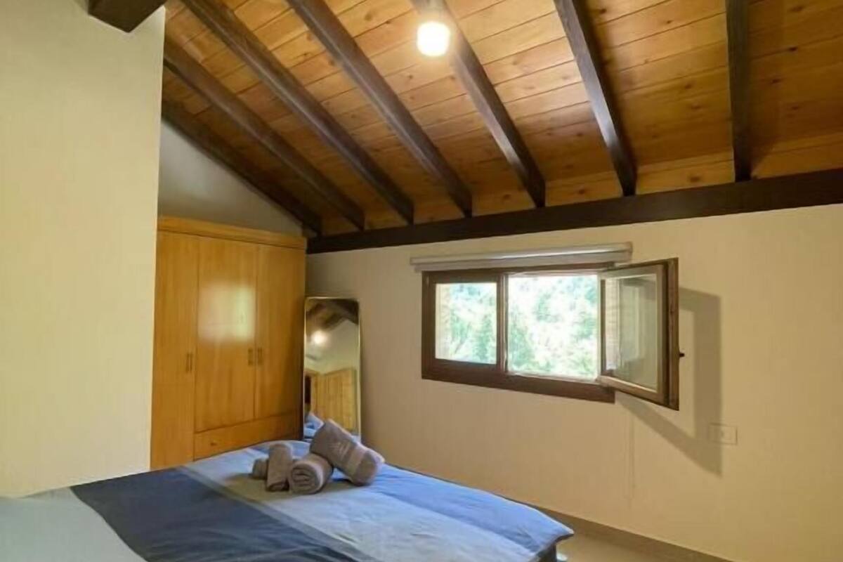 A bedroom with a wooden ceiling, a bed with a blue and white mattress, and a window with a view of greenery.