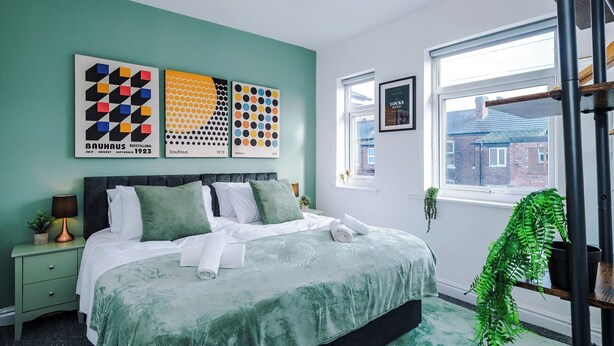 Un dormitorio moderno con una cama, paredes verdes, obras de arte, una ventana y una planta.