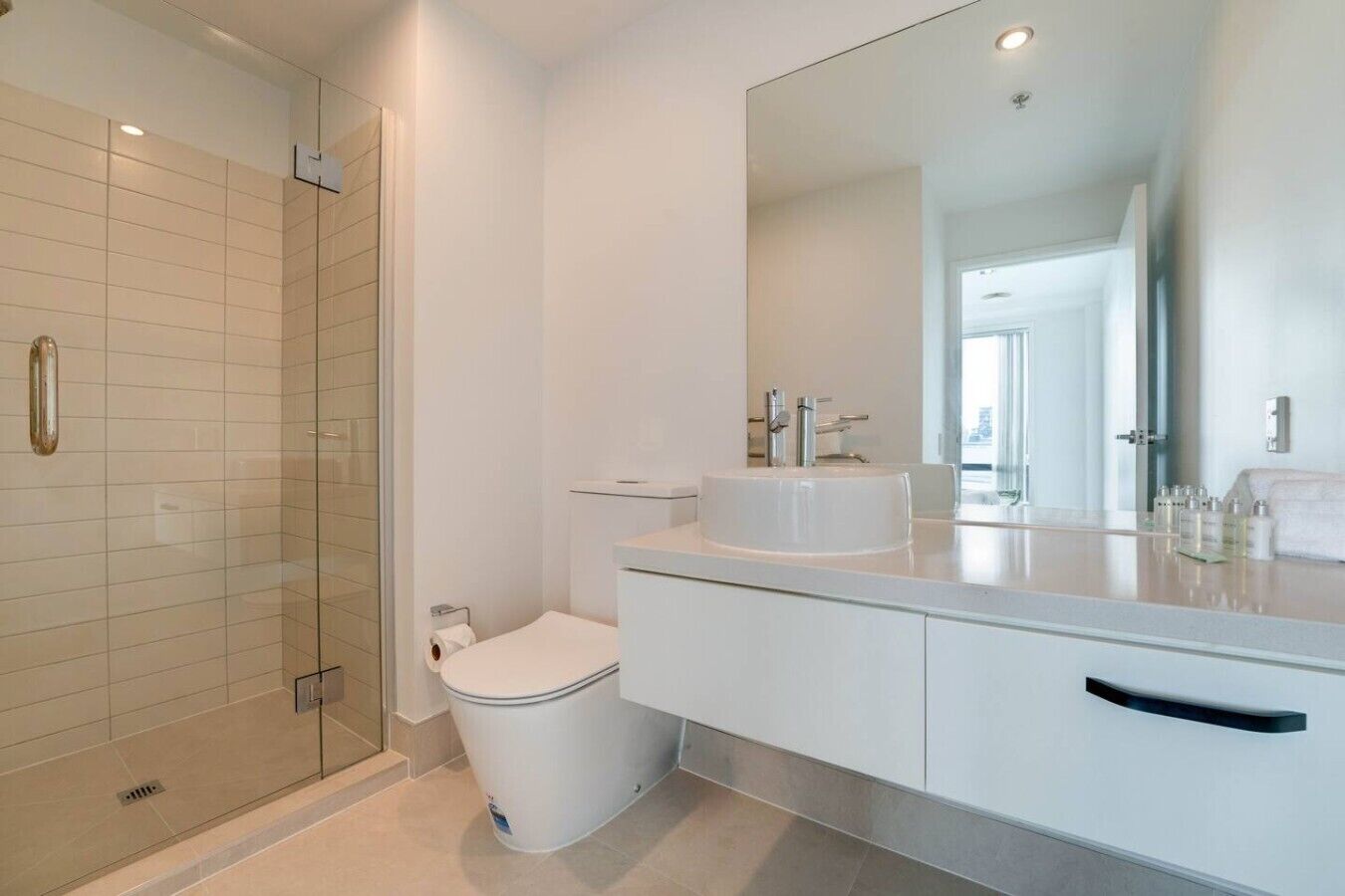 Ein modernes Badezimmer mit Duschkabine aus Glas, einer weißen Toilette und einem großen Spiegel über einem weißen Waschbecken mit Arbeitsplatte.