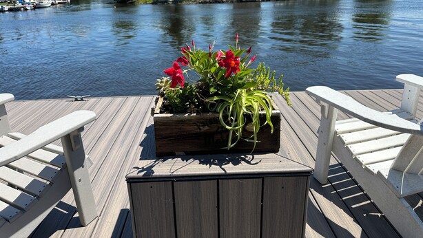 Un muelle de madera con una jardinera con flores rojas, una pequeña mesa y sillas blancas.