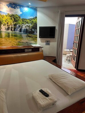 A hotel room with a bed, a remote control, a wall-mounted air conditioning unit, and a scenic mural of a waterfall. - Johnnys Motel-Hotel (Cornélio Procópio)