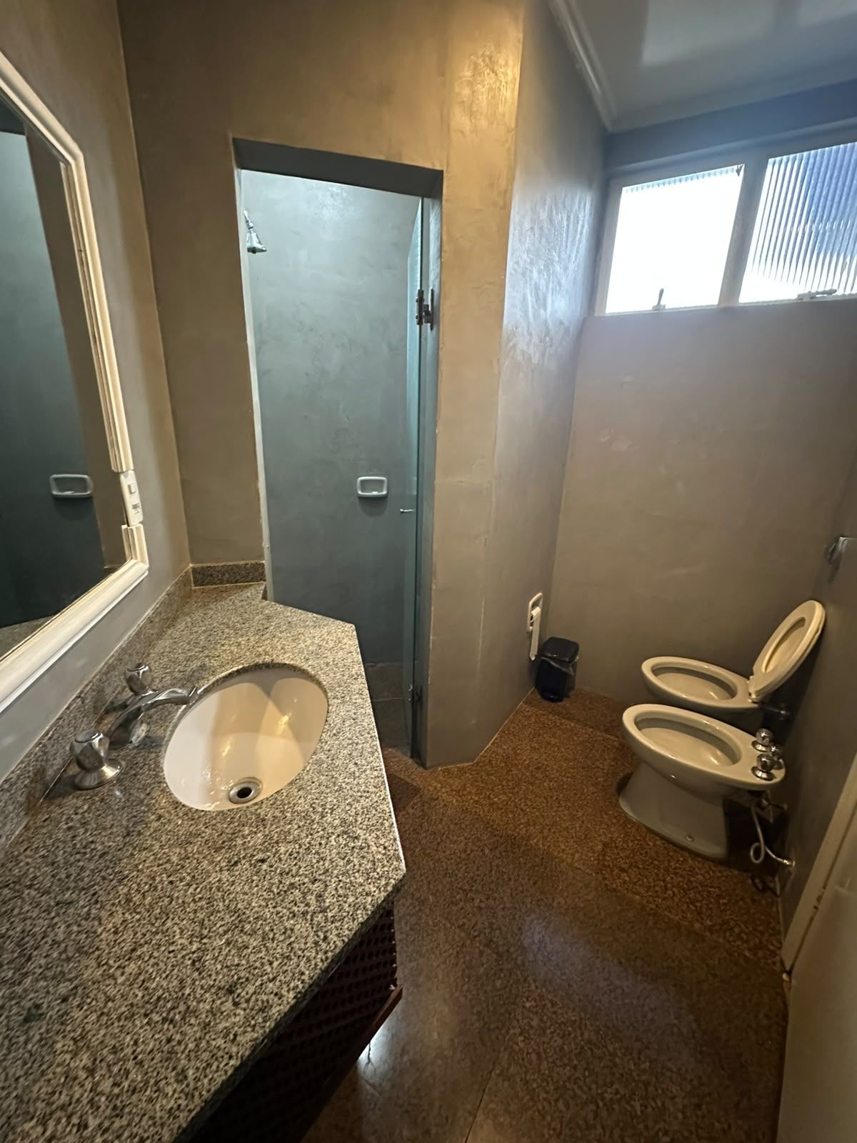 Un baño con lavamanos de encimera de granito, un espejo, una puerta que da a otra habitación y dos inodoros.