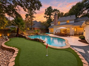 A backyard with a swimming pool, a putting green, and a patio area with outdoor furniture.