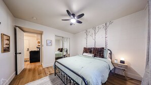 A bedroom with a bed, a nightstand, a ceiling fan, a mirror, and a door leading to another room.
