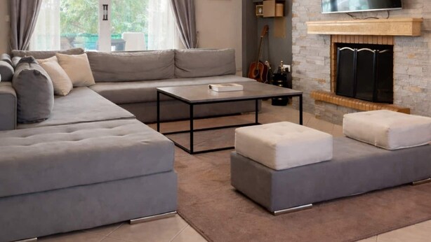 Un salon moderne comprenant un canapé d’angle, une table basse, des poufs, une cheminée, un téléviseur fixé au mur et une grande fenêtre avec des rideaux.
