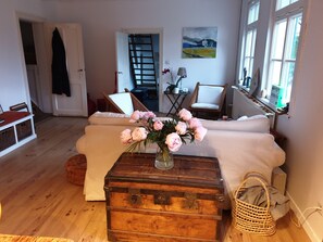 Een woonkamer met een houten kist, een bank, een stoel, een bijzettafeltje met een bloevas, en een schilderij aan de muur.