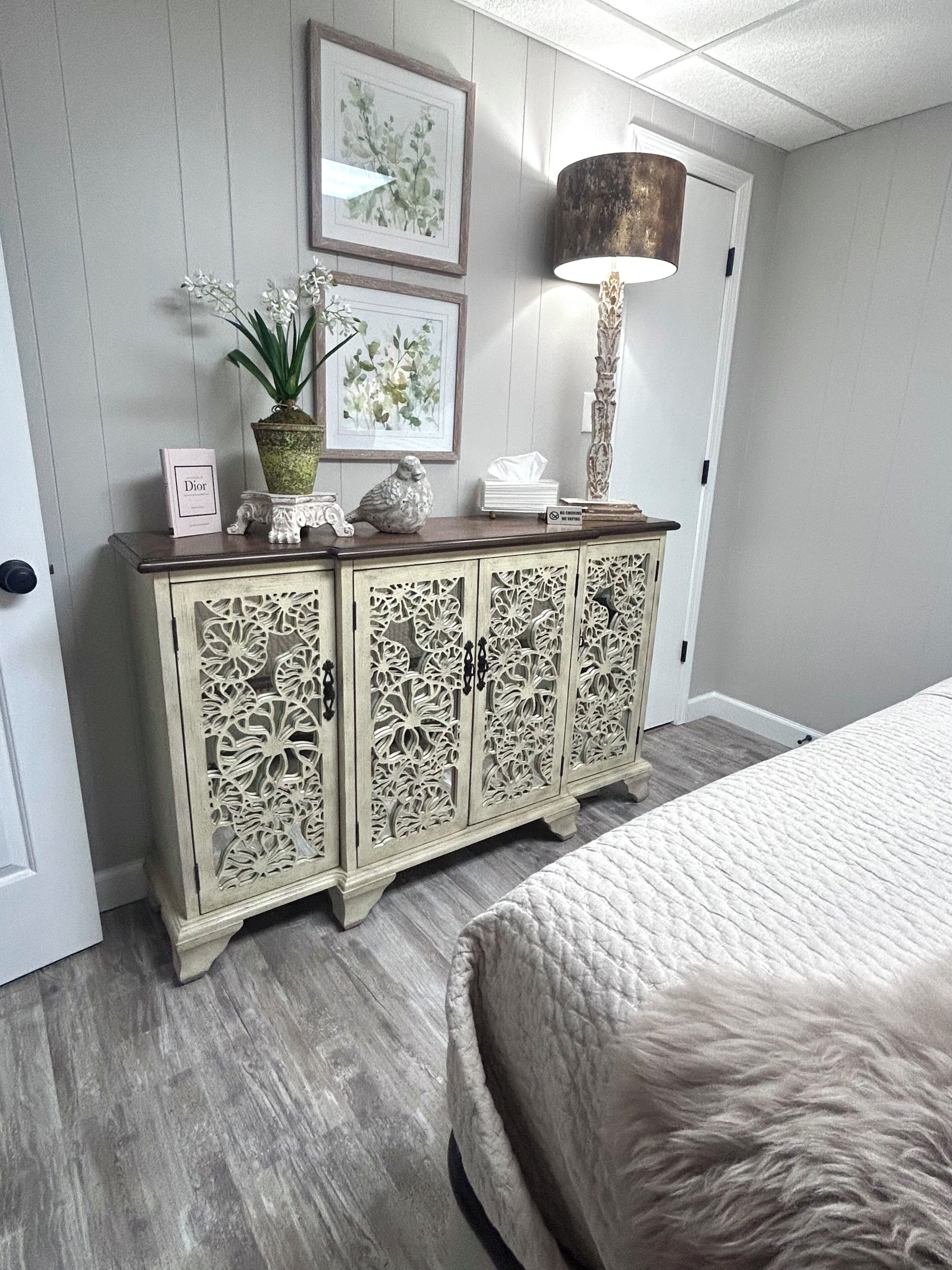 A bedroom with a bed, a cabinet with intricate carvings, a lamp, and framed artwork on the wall.