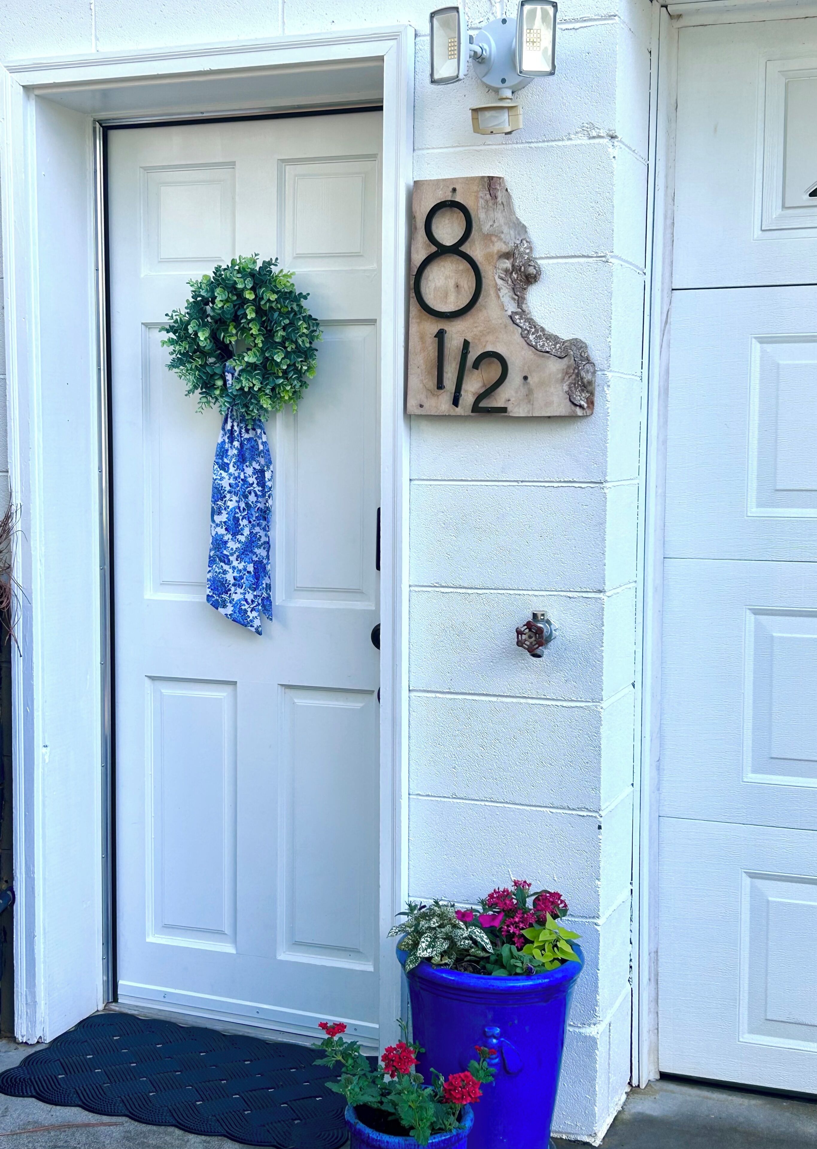 Une porte blanche ornée d’une guirlande, une boîte aux lettres en bois portant le numéro 8 1/2, et un pot de fleurs bleu.