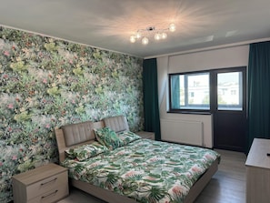 A bedroom with a floral wallpaper, a bed with a matching floral bedspread, a nightstand, a dresser, and a window with curtains.
