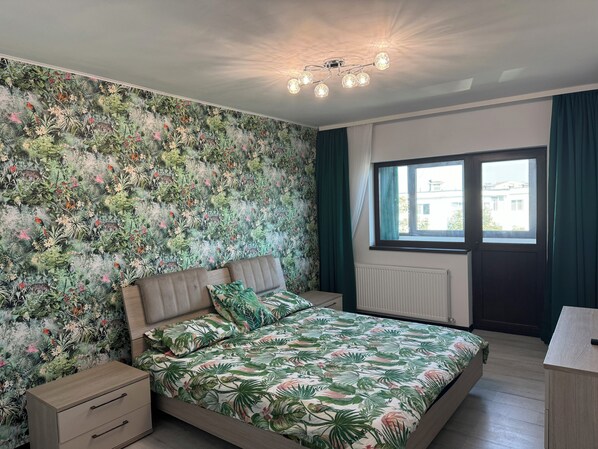 Un dormitorio con papel tapiz floral, una cama con colcha floral a juego, una mesita de noche, un aparador y una ventana con cortinas.