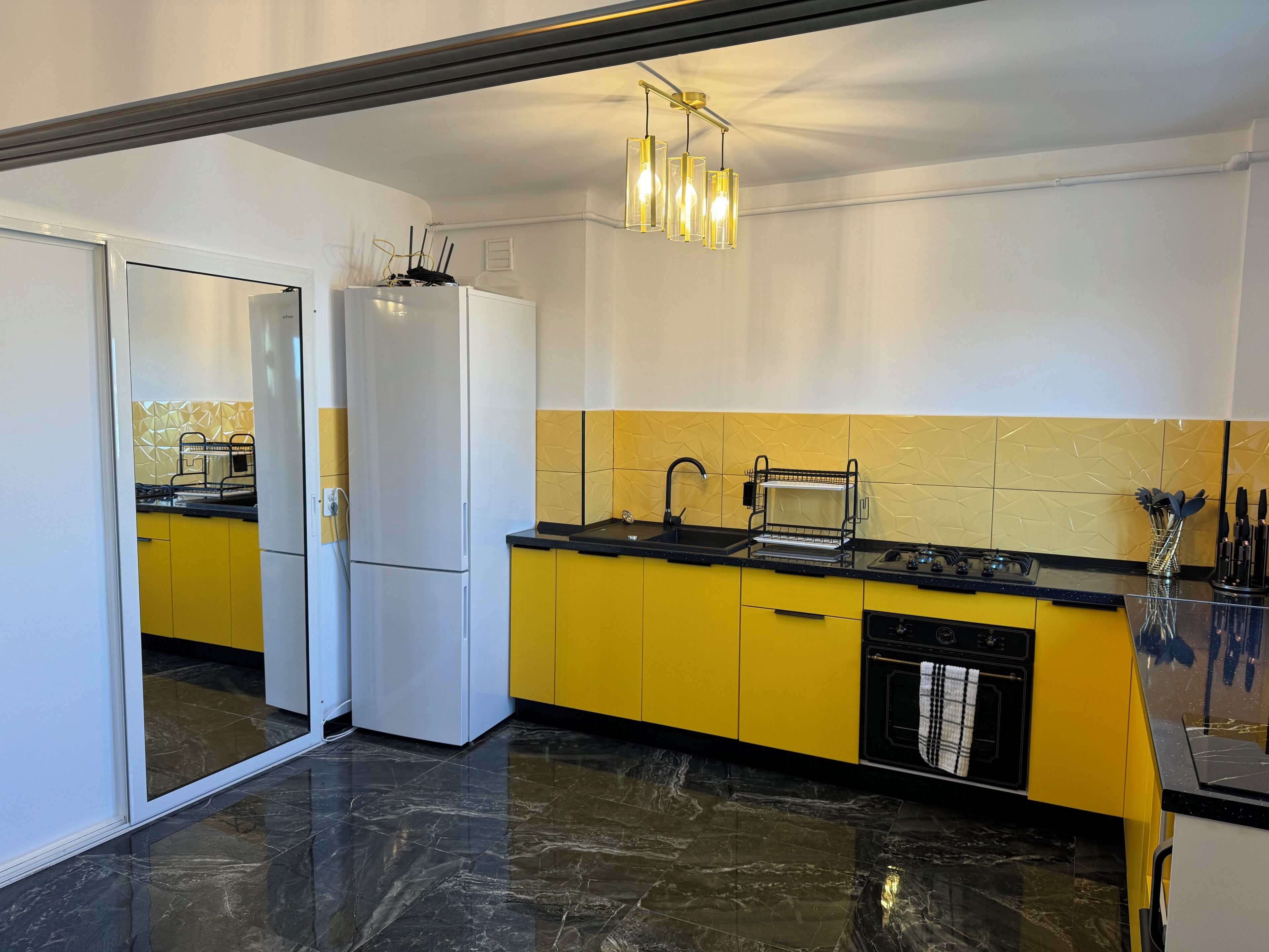 A modern kitchen with a white refrigerator, yellow cabinets, a black countertop, a sink, and a hanging light fixture.