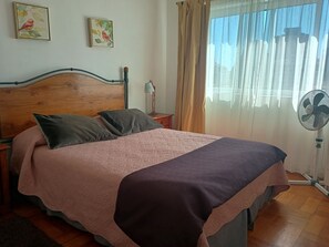 Ein Bett mit Holzkopfseitenverkleidung, zwei Kissen, ein Nachttisch mit Lampe, ein Fenster mit Vorhängen und ein Standventilator.