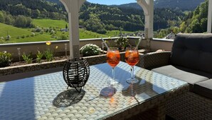 Un tavolo con piano in vetro, due bicchieri con una bevanda, una sedia in vimini e vista su un paesaggio verde.
