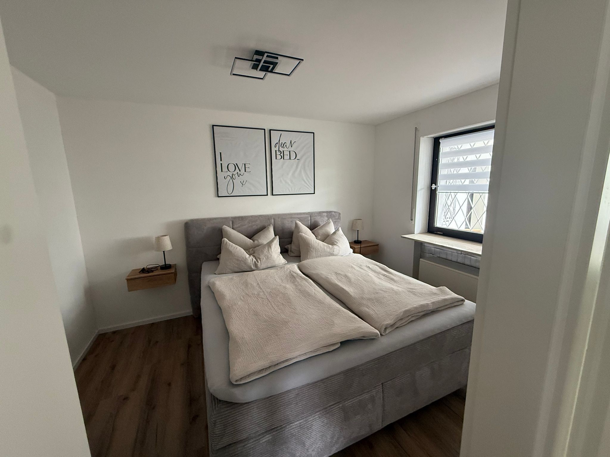 Un dormitorio con una cama, mesitas de noche, una ventana y dos cuadros en la pared.